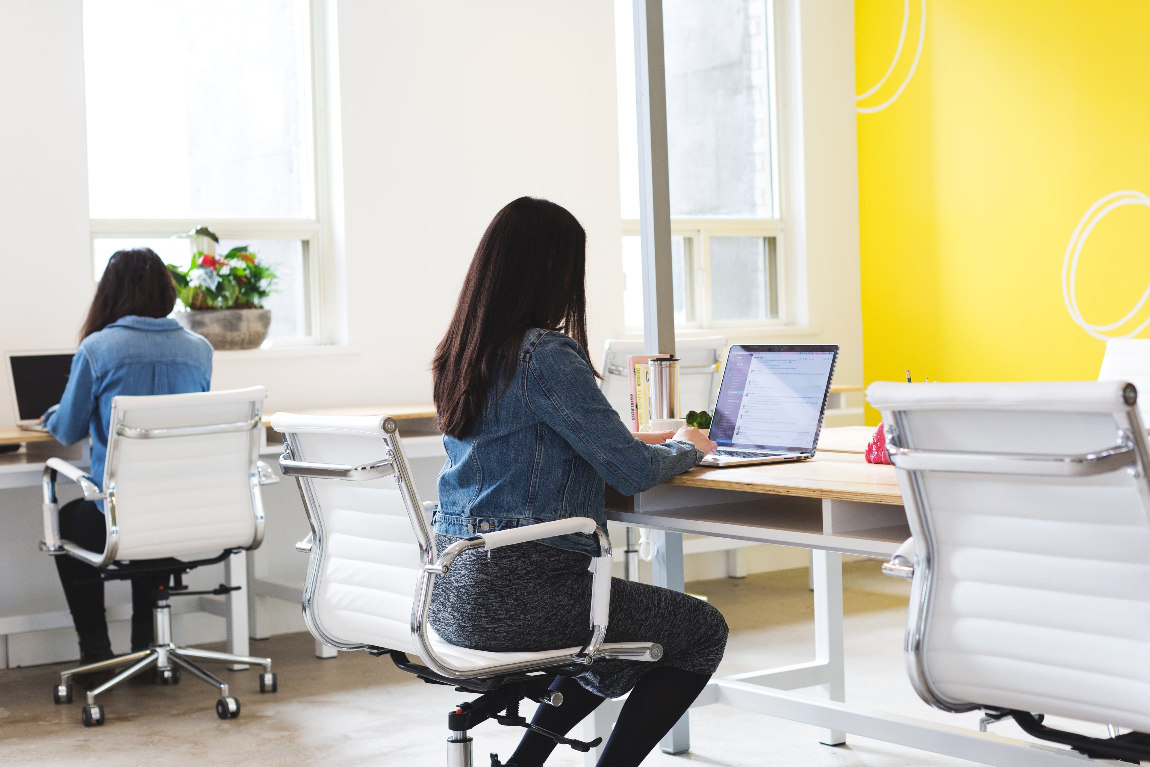Two women sit at separate desks in a bright, modern office with white chairs and a yellow accent wall. One works on a laptop, while the other faces away, using a computer. Natural light fills the room.